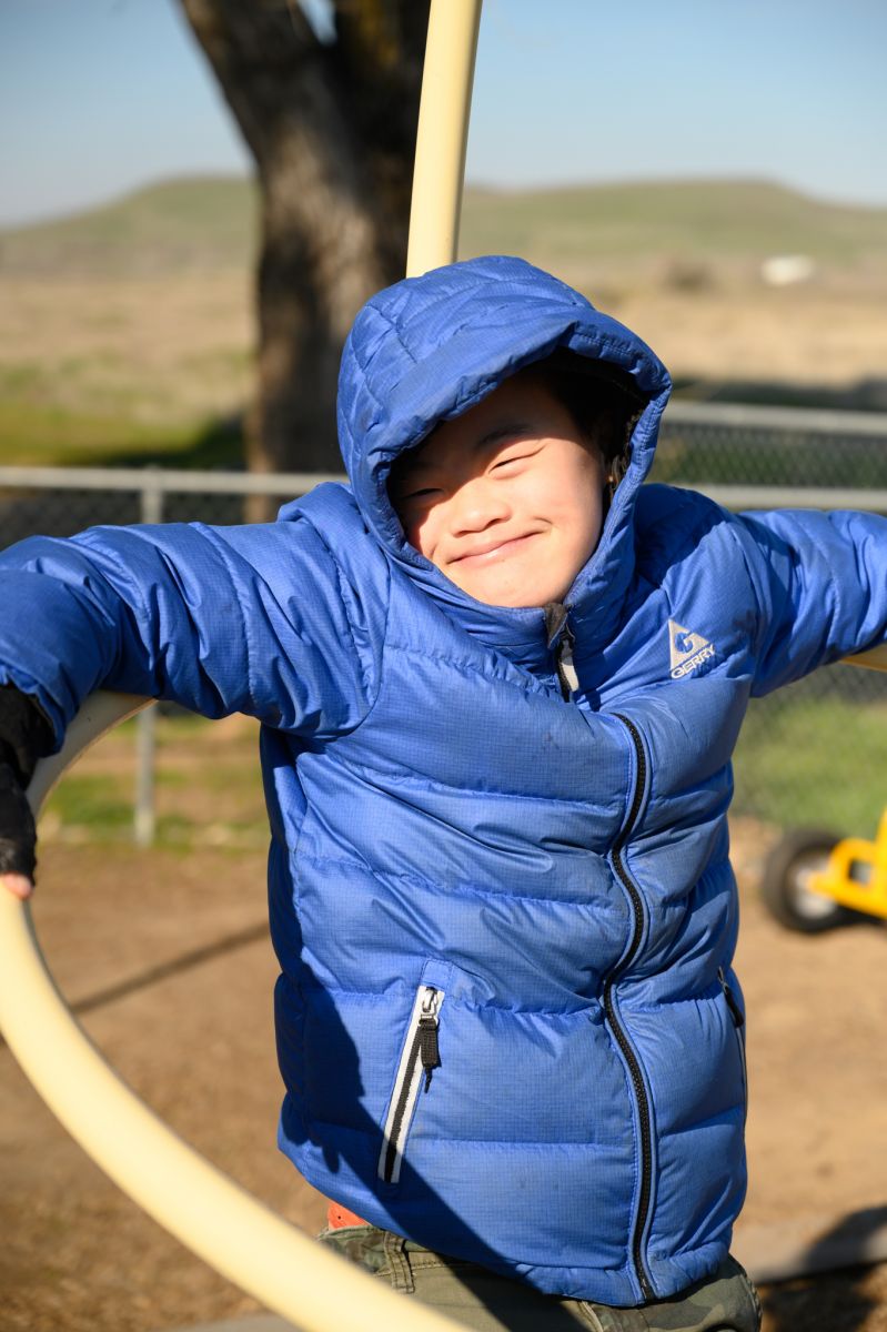 Student smiling for the camera in a blue quilted jacket with a hood, while sitting on play equipment.